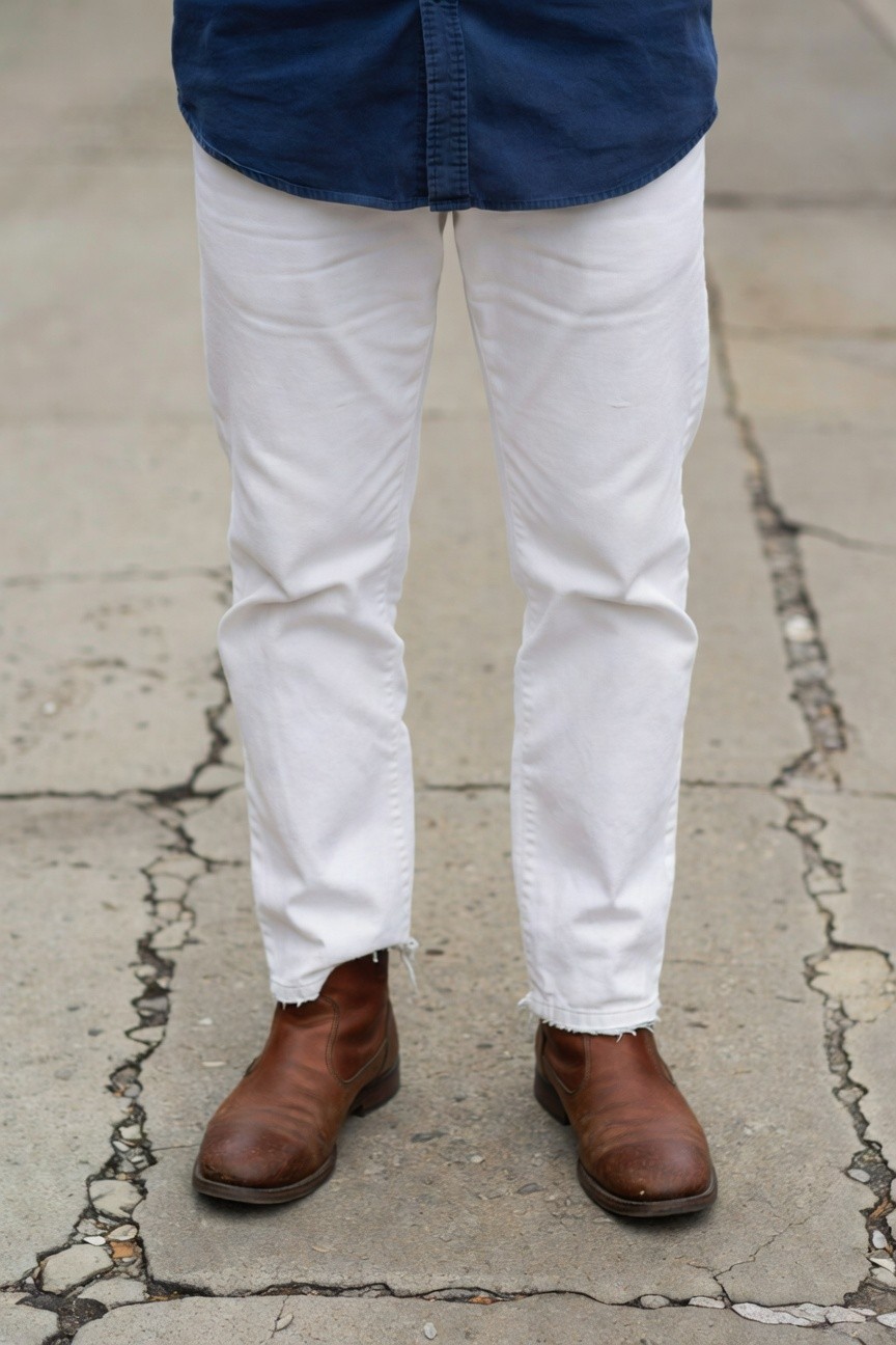 Man in untucked blue chambray shirt over slim white cuffed pants and brown leather ankle boots standing on cracked urban pavement