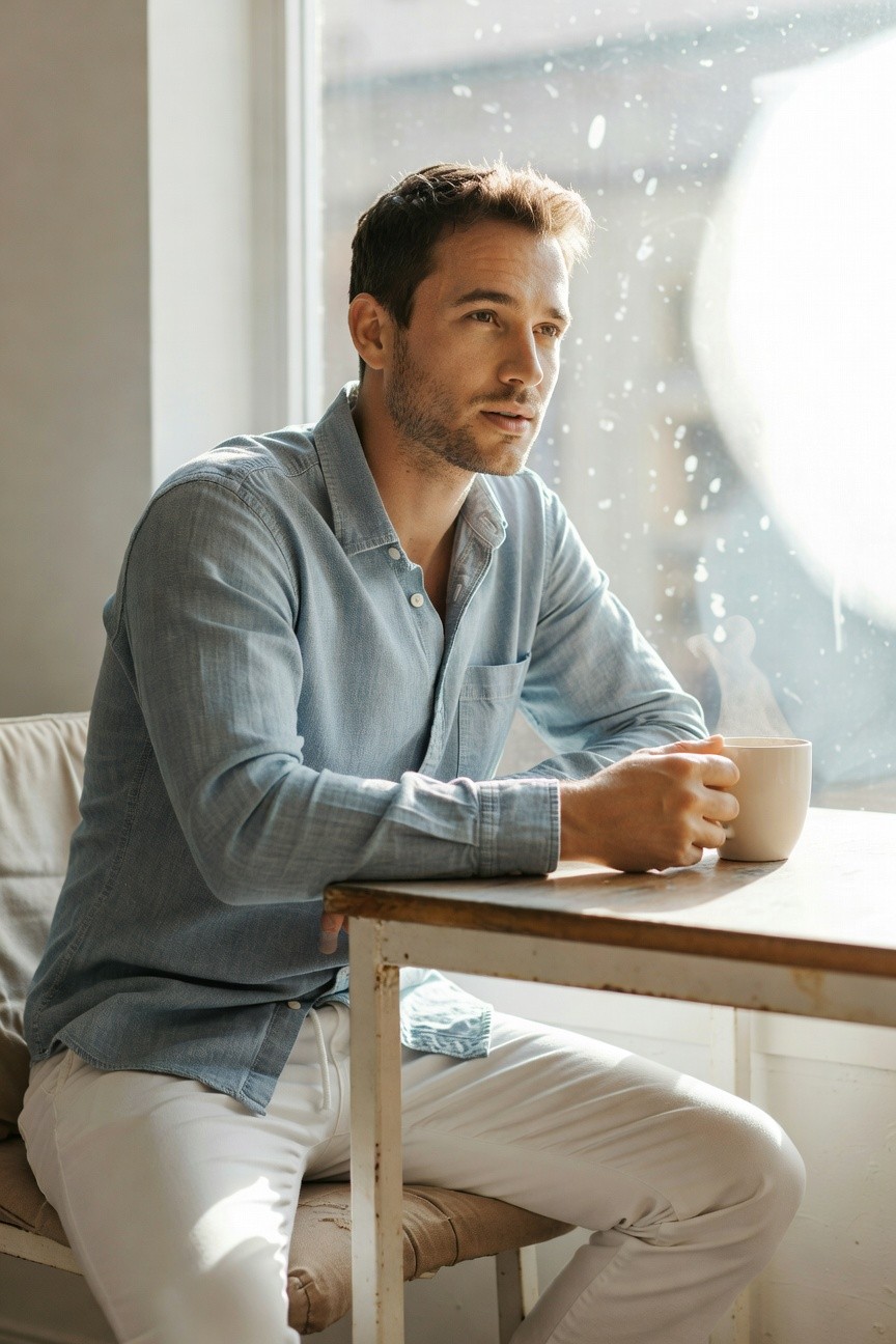 A man with short brown hair sits pensively by a sunlit window in a cafe, wearing a light blue linen long-sleeve shirt with top buttons undone, slim white linen pants, holding a ceramic mug of coffee on a wooden table with a white chair beneath him