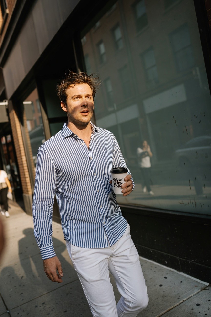 A stylish man in a light blue vertically striped long-sleeve button-up shirt with an open collar, white slim trousers, holding a white lidded coffee cup, walking casually on a sunny urban sidewalk beside large glass building windows with blurred passersby in background.