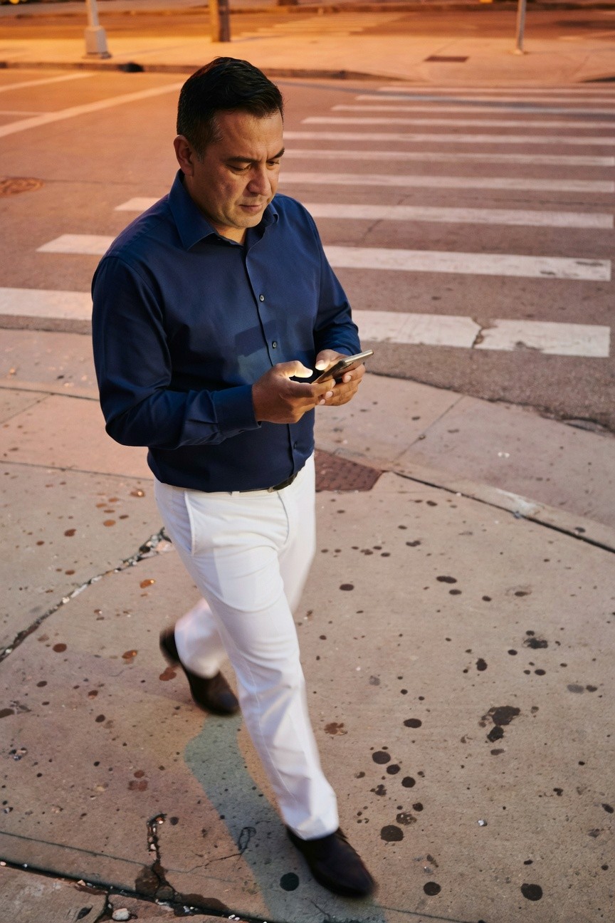 Man in navy blue long-sleeve button-up shirt and slim white trousers walking on urban crosswalk at dusk, checking phone, black shoes, streetlights glowing