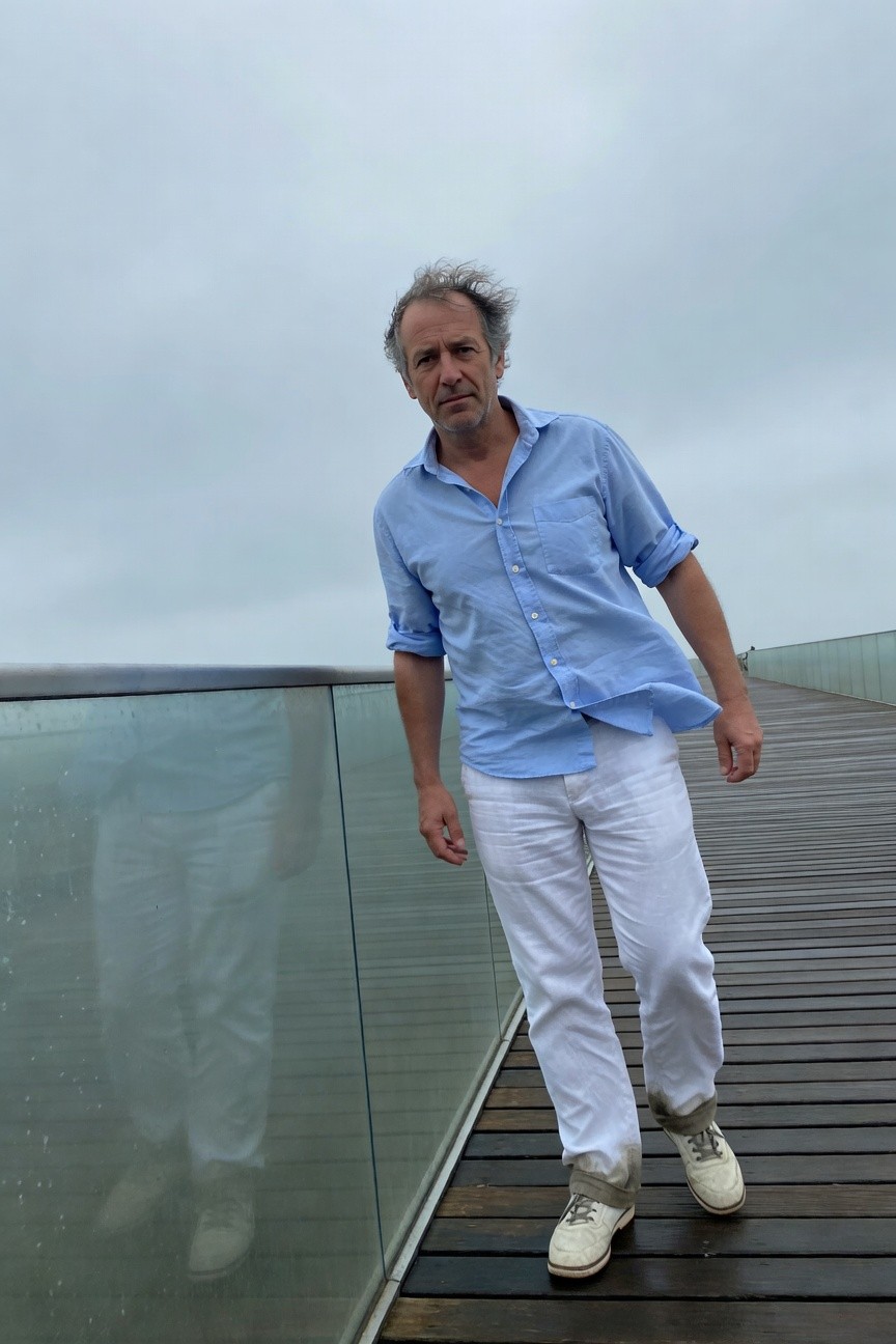 Middle-aged man in light blue button-up shirt with rolled sleeves and white linen pants stands casually on a wooden pier with glass railing over gray water under cloudy skies wearing white sneakers
