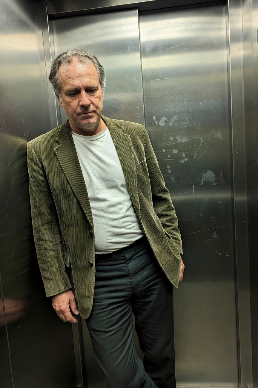 Man in olive corduroy blazer over white t-shirt, charcoal gray trousers, brown belt and shoes, leaning casually in a metallic elevator with hands in pockets
