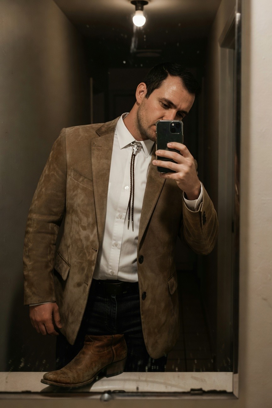 Man taking mirror selfie in brown suede blazer, white dress shirt, bolo tie, dark pants, and brown cowboy boots for a modern cowboy wedding look
