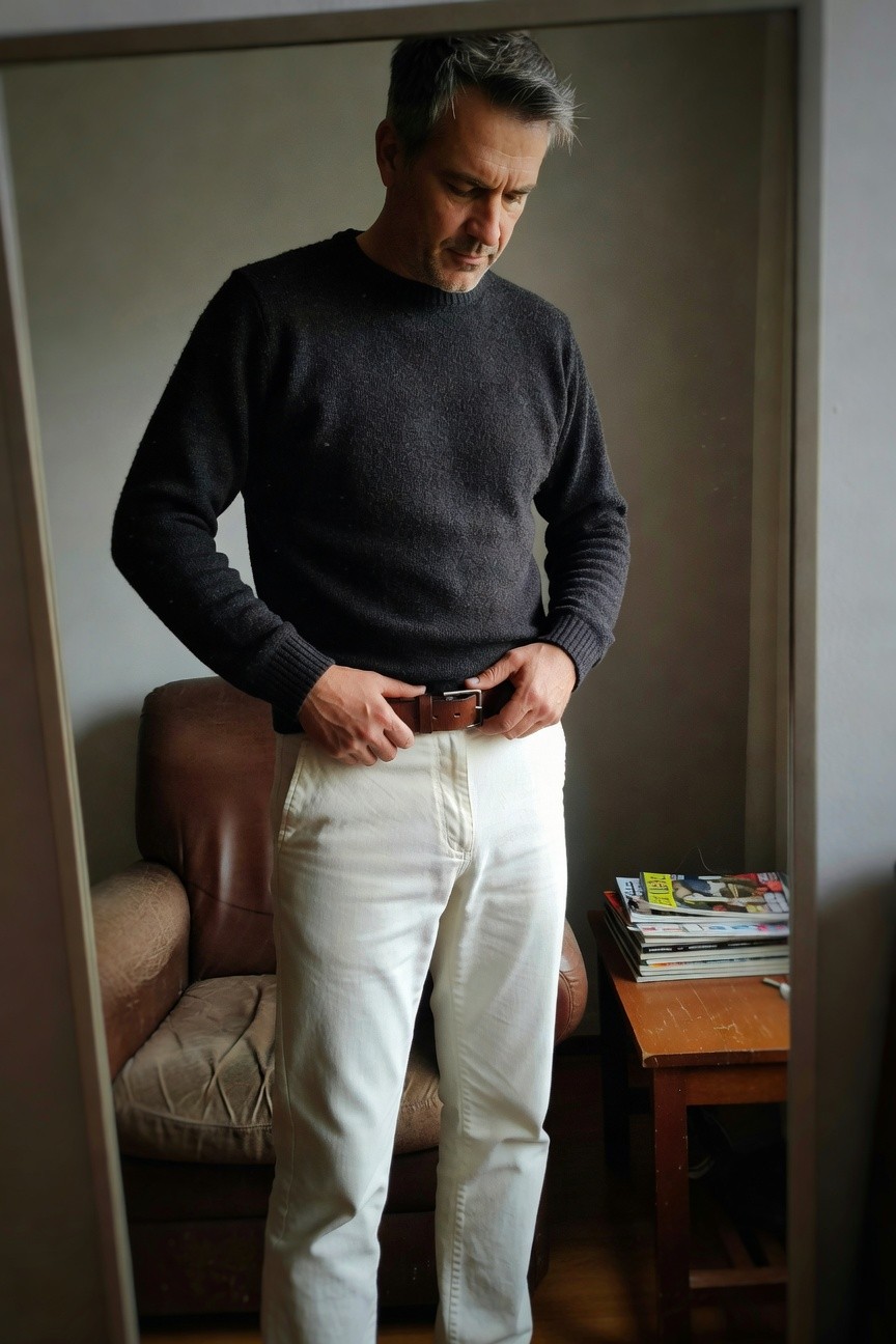 Silver-haired man in charcoal crewneck sweater, white trousers, and brown leather belt stands confidently before a full-length mirror in a warmly lit room with leather armchair and stacked magazines nearby