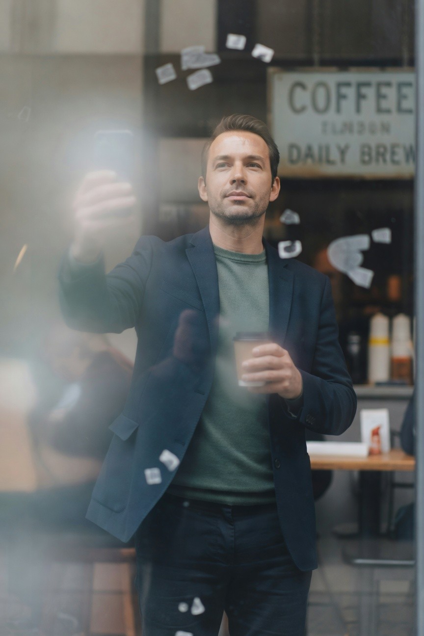 A stylish man in a navy blazer over a green t-shirt and dark trousers stands outside a coffee shop window, holding a coffee cup in one hand and taking a selfie with his phone in the other, with blurred interior and floating paper bits visible through the glass