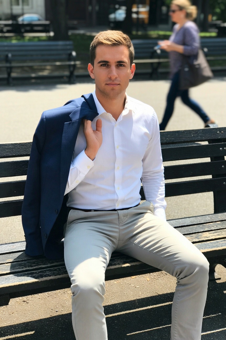 A young man with short blond hair sits on a black park bench wearing a navy blue blazer draped over one shoulder a crisp white button-up shirt tucked into light gray slim-fit trousers and a black belt urban park setting with trees benches and passersby in background
