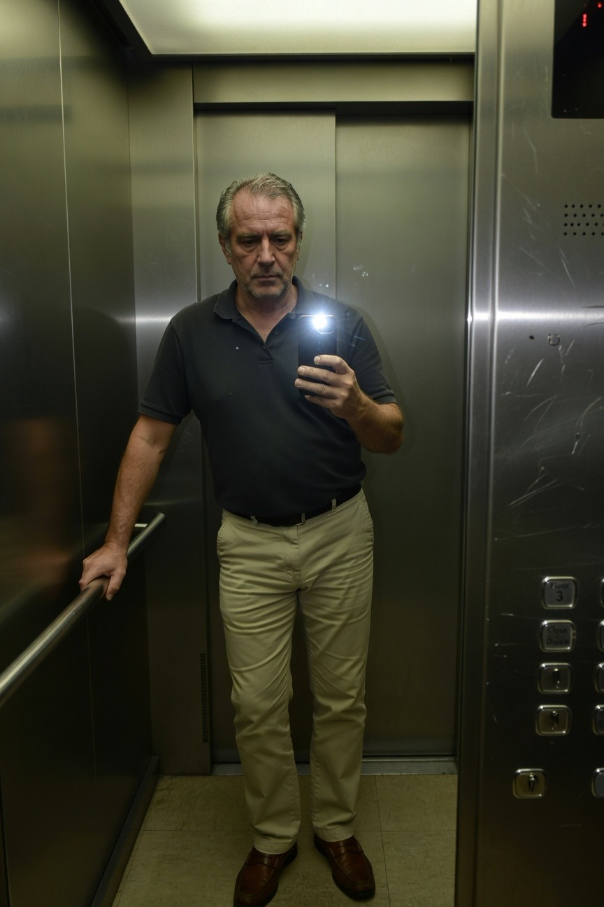 Middle-aged man in black short-sleeve polo shirt, khaki chinos, and brown loafers, taking a selfie with phone flashlight on inside a steel elevator, casual professional summer work attire