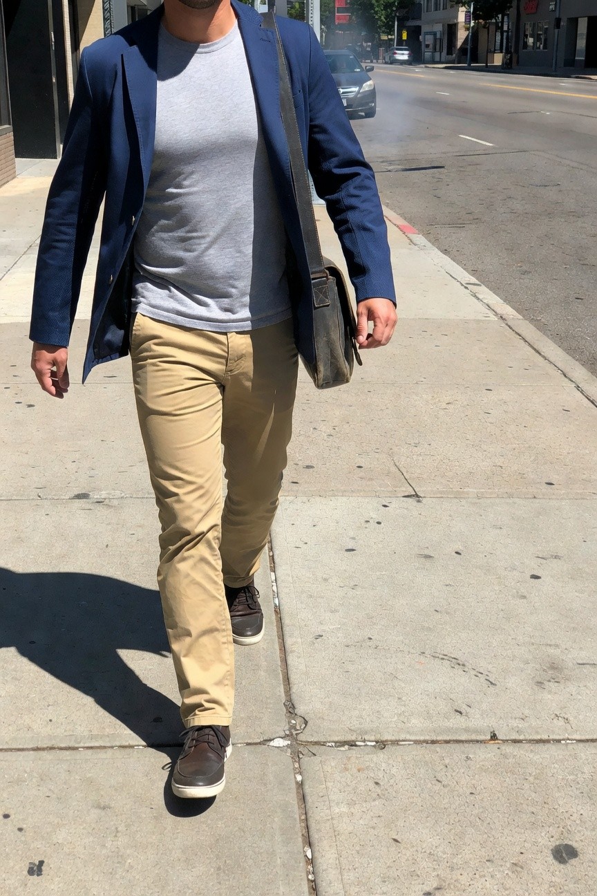 A man walks on a sunny urban sidewalk wearing an open navy blue blazer over a gray t-shirt, beige khaki pants, brown leather sneakers, and carrying a tan messenger bag, evoking casual summer work style.