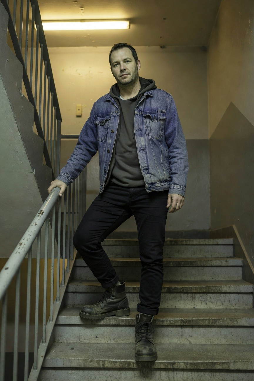 Man leaning on stair railing in a dimly lit stairwell, dressed in a faded blue denim jacket over a gray hoodie, slim black jeans, and black combat boots, exuding relaxed urban style