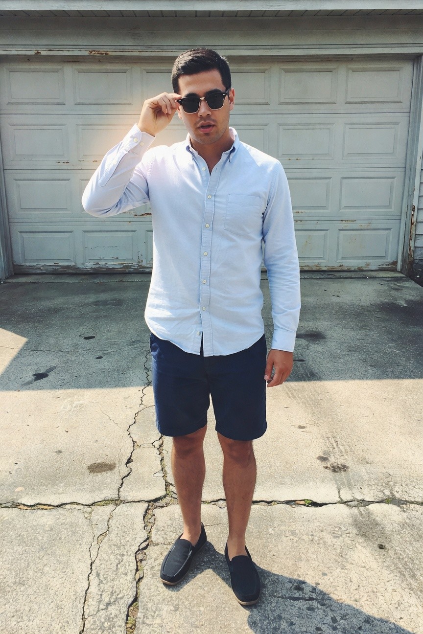 Man in light blue chambray button-down shirt with rolled sleeves, navy tailored shorts, black suede loafers without socks, and sunglasses, posing casually in front of a white garage door on a concrete driveway