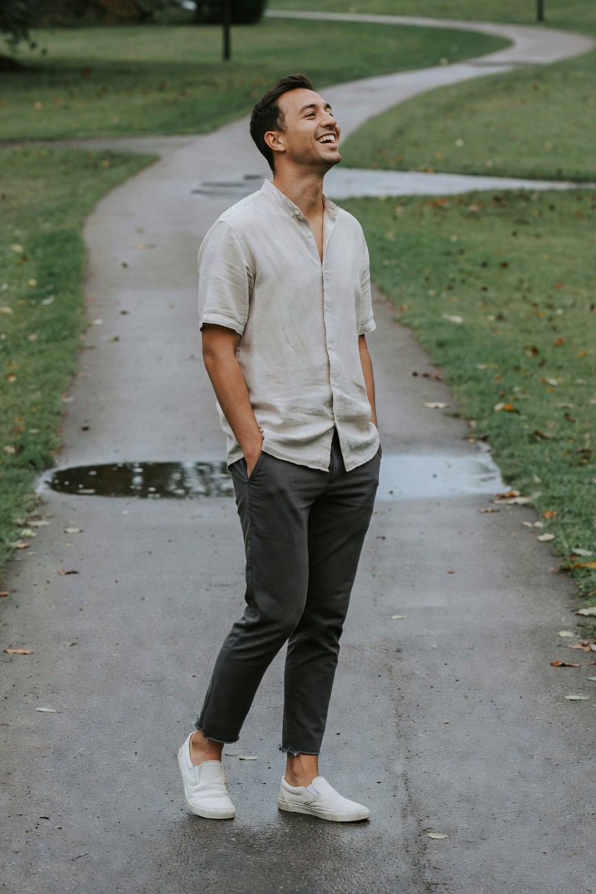 A smiling man in a park path after rain, wearing an open pale linen short-sleeve shirt, slim grey cropped pants, and white sneakers, hands in pockets for a relaxed polished casual look.