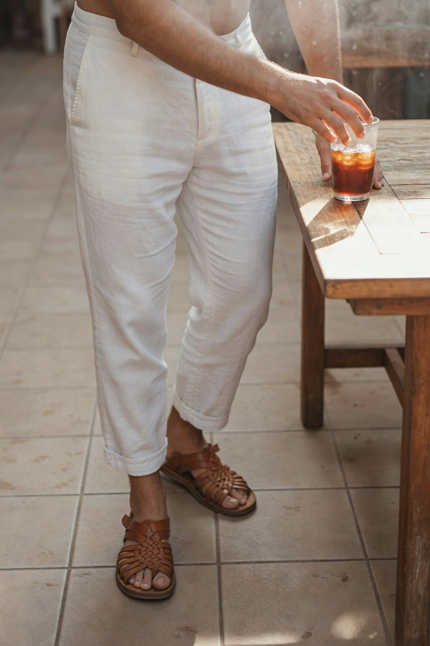 A man stands relaxed on a sunlit patio in wide-leg white linen trousers cuffed at the ankles, brown woven leather sandals, holding an iced tea glass, shirtless with casual vibe.