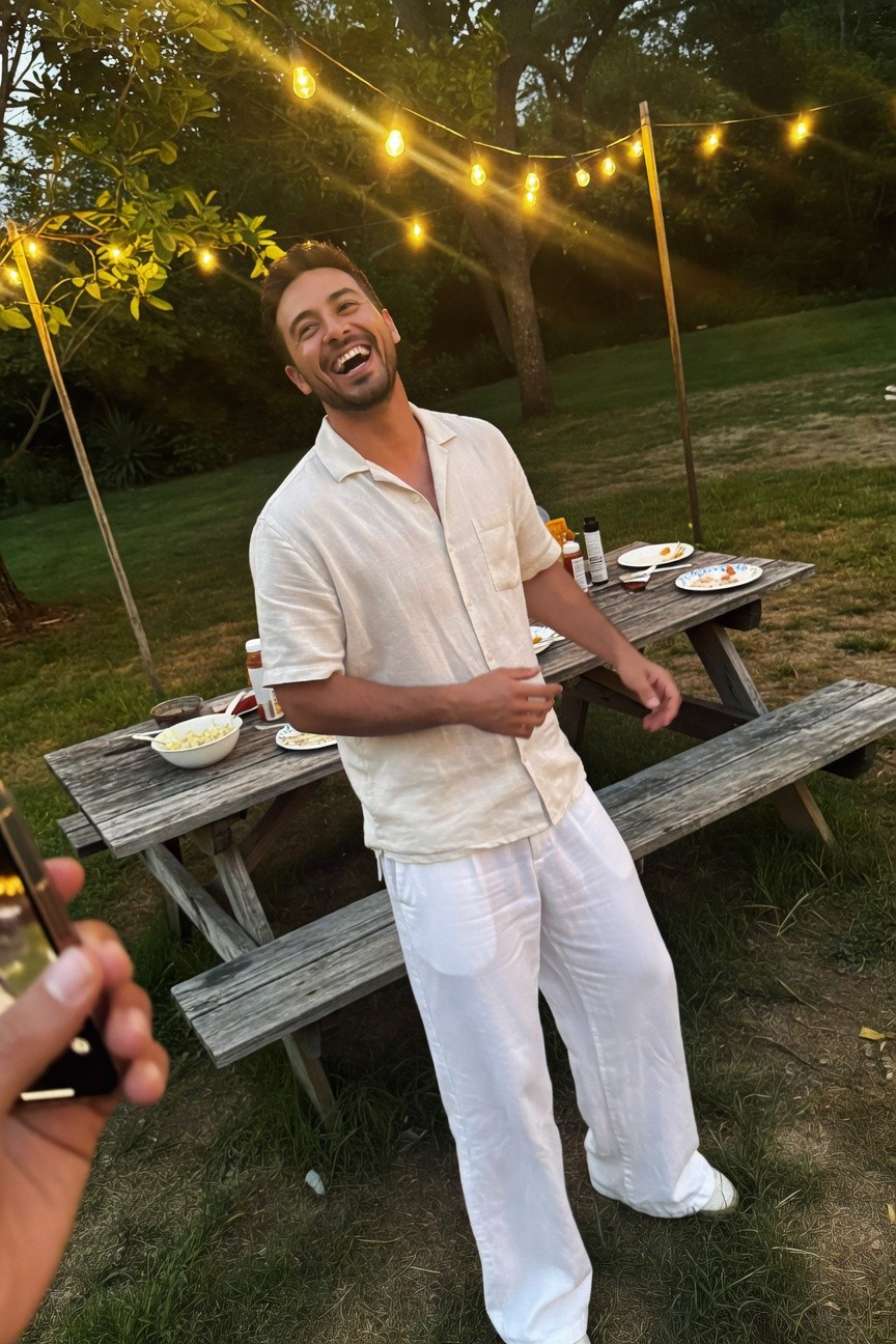 Smiling man in open white linen shirt and matching wide-leg pants dances lightly by outdoor picnic table at dusk with string lights and food spread