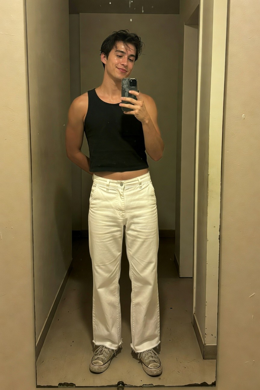 Young man taking a mirror selfie in a beige hallway, wearing a fitted black sleeveless tank top exposing midriff slightly, high-waisted white wide-leg pants, and worn gray sneakers, arms relaxed at sides for a casual confident pose