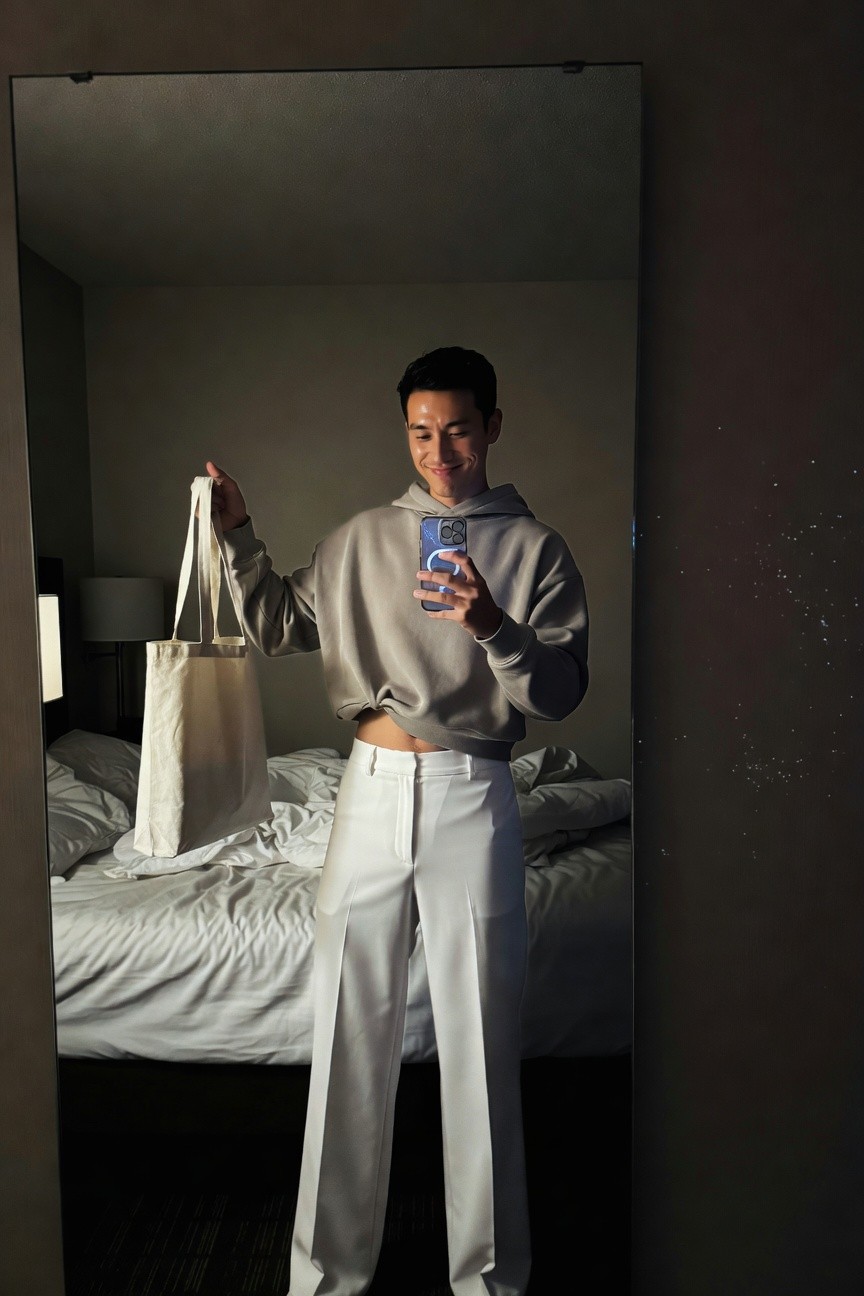 Young Asian man in cropped gray hoodie revealing midriff, high-waisted white wide-leg pants, holding beige tote bag, mirror selfie in hotel room with bed visible