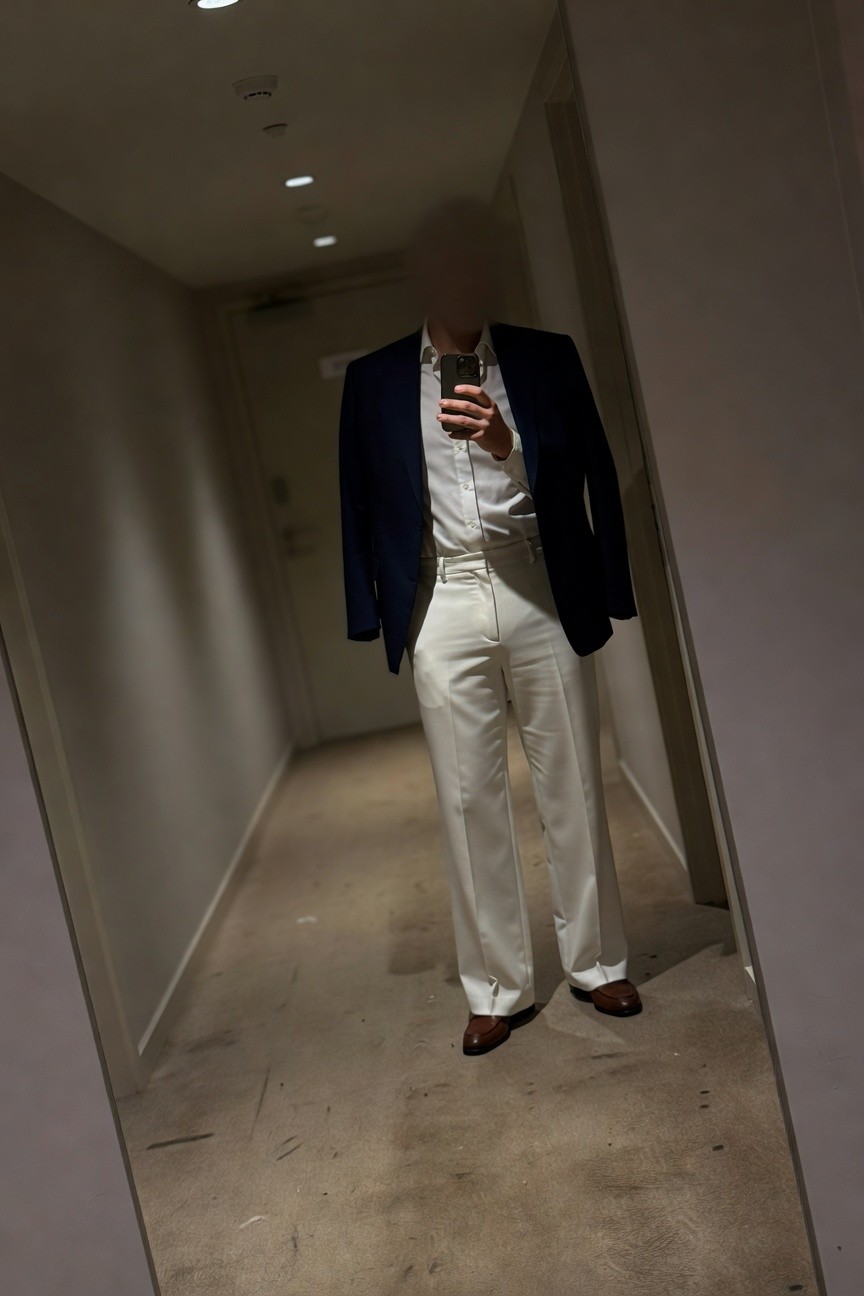 Man in navy blazer, white dress shirt, and flowing white wide-leg pants stands confidently in a dimly lit hotel hallway mirror selfie, brown dress shoes grounding the sophisticated ensemble