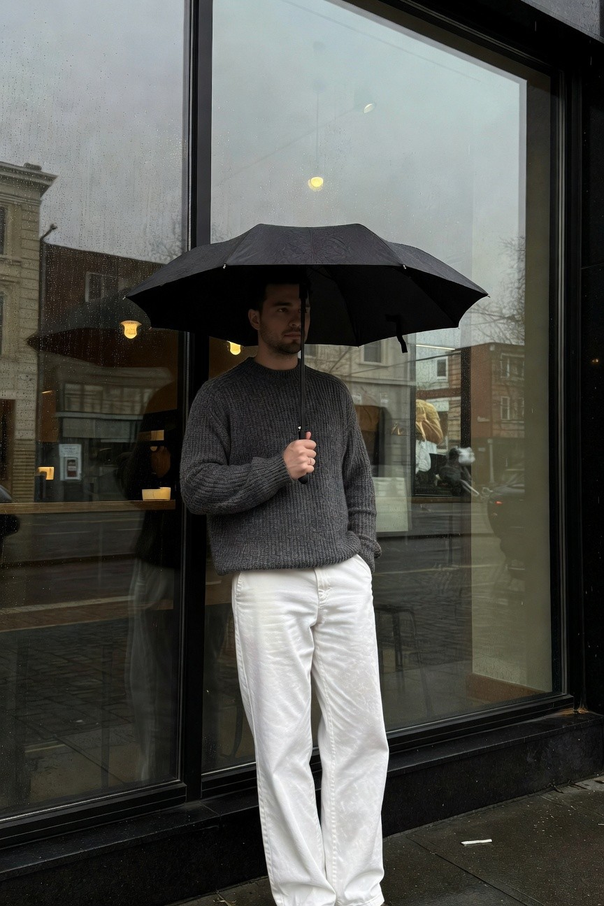 Man in chunky gray sweater and crisp white wide-leg pants stands confidently outside a rainy storefront window holding a large black umbrella with wet pavement reflecting blurred lights