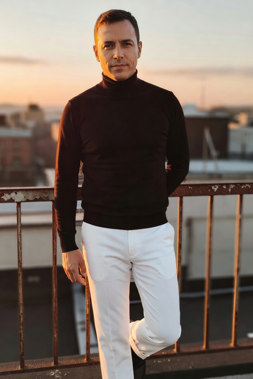 Confident man in fitted black turtleneck sweater and slim white trousers leans against rusted rooftop railing at sunset, cityscape and orange sky in background