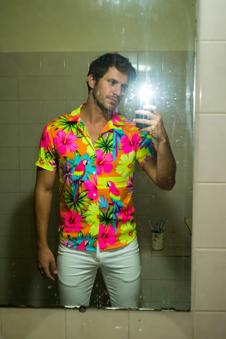 A man takes a mirror selfie in a steamy bathroom, wearing a short-sleeved floral Hawaiian shirt in vivid pinks, yellows, blues, and greens with hibiscus patterns, paired with form-fitting white pants; he holds his phone with a slight smile, beard and tousled hair adding casual vibe.