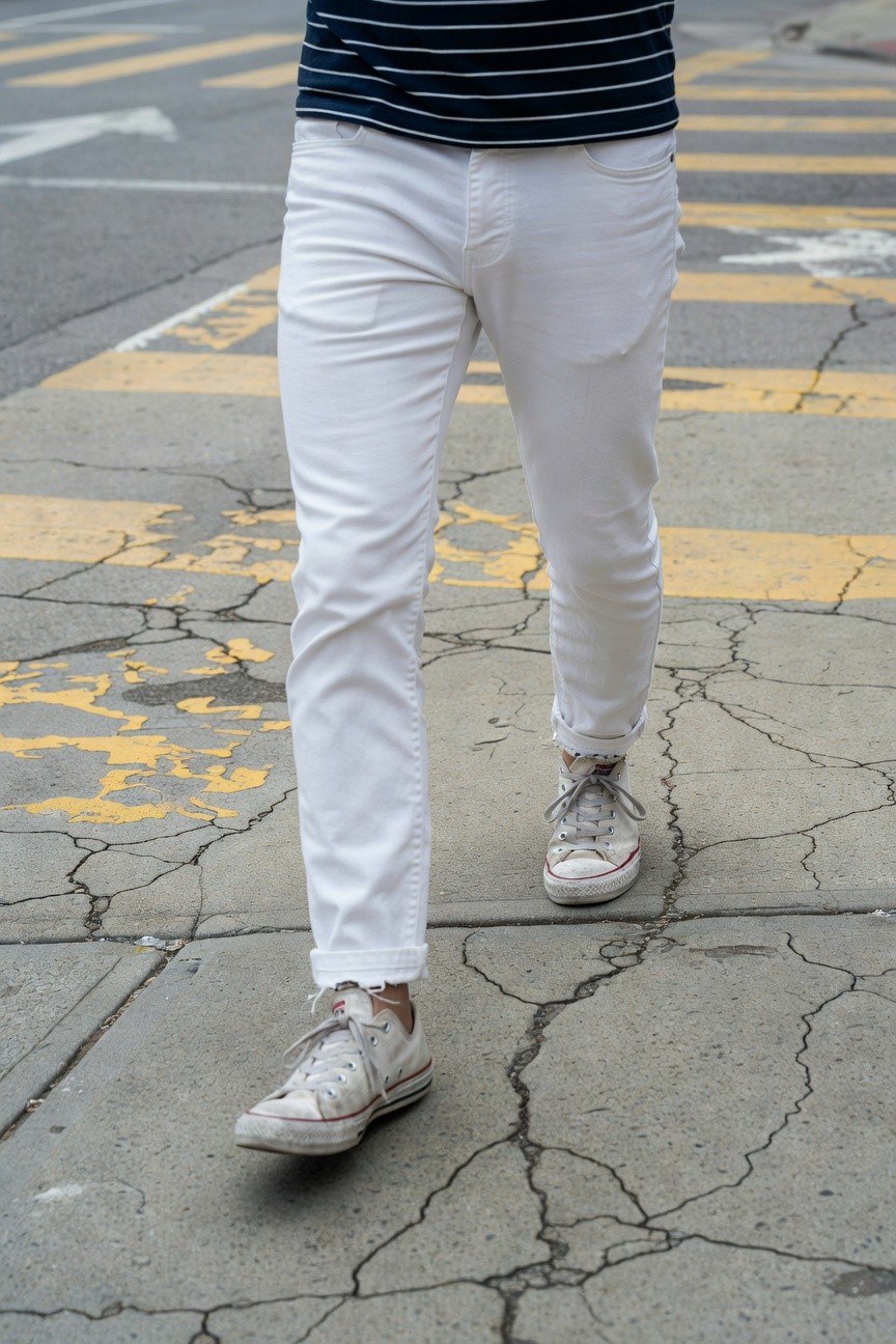 A man walking on a cracked urban street crossing, wearing a navy and white horizontal striped short-sleeve shirt tucked into slim white pants cuffed at the ankles, paired with white low-top Converse sneakers, against a background of yellow pedestrian stripes and asphalt.