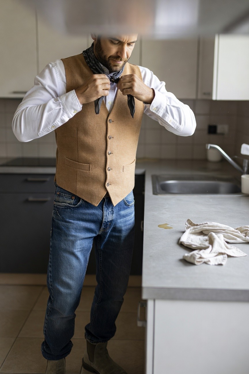 Man adjusting black neckerchief under open tan vest over white shirt, wearing slim blue jeans and tan boots while standing in modern kitchen