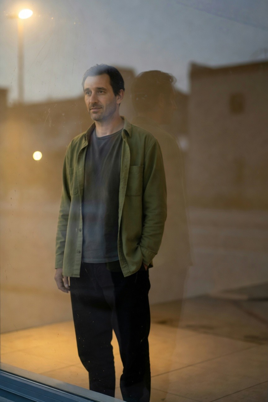 Man in olive green shacket over black t-shirt and black pants stands pensively by a nighttime window, city lights reflecting softly.