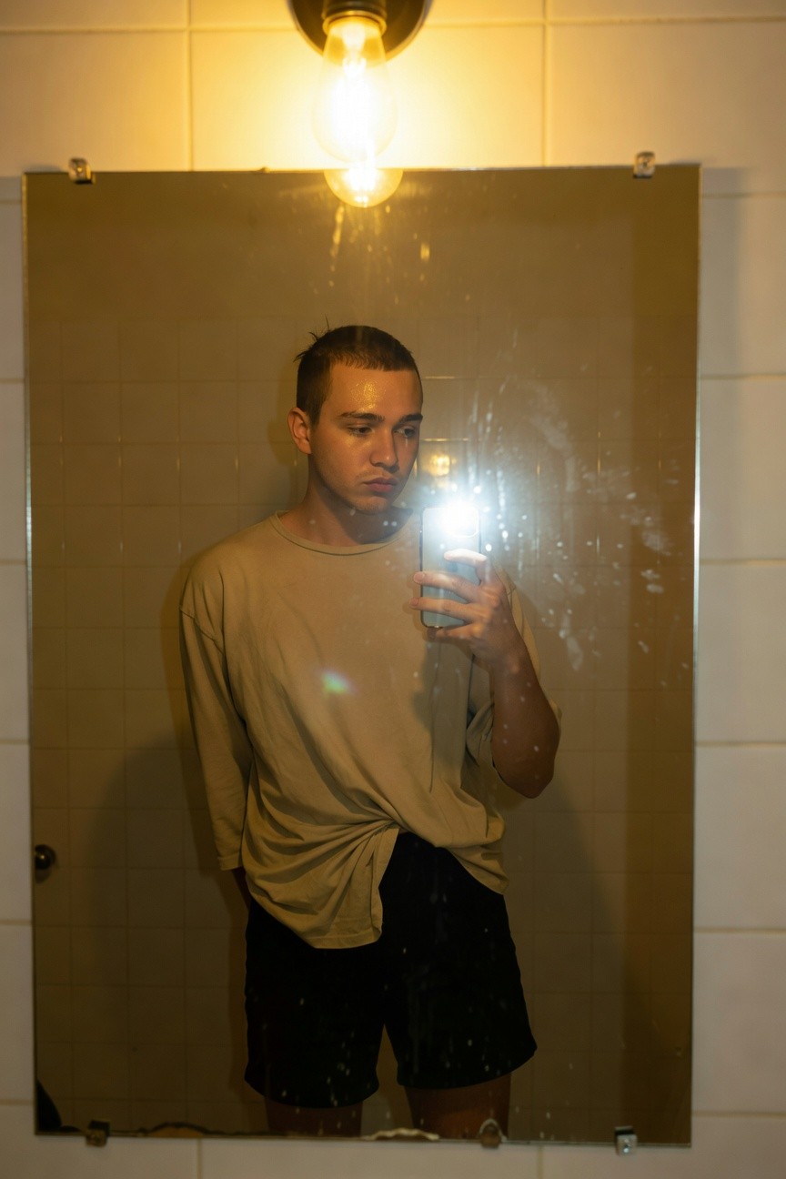 Young man in a steamy bathroom mirror selfie wearing a loose light beige oversized t-shirt casually twisted at the waist tucked loosely over black mid-thigh shorts standing relaxed with neutral expression under warm wall light