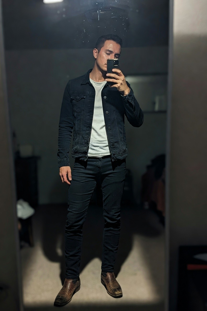 Man in mirror selfie wearing dark denim jacket over white t-shirt, slim black jeans, and brown leather boots for a modern cowboy style