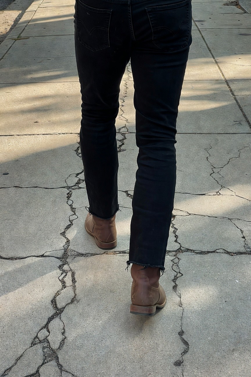 Rear view of a man walking in slim black frayed-hem jeans over tan leather ankle boots on a cracked sidewalk