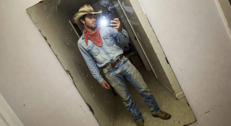 Man in beige cowboy hat, red bandana neckerchief, long-sleeve blue denim shirt, distressed blue jeans, brown belt, and brown cowboy boots, posing in front of a wooden mirror for a selfie
