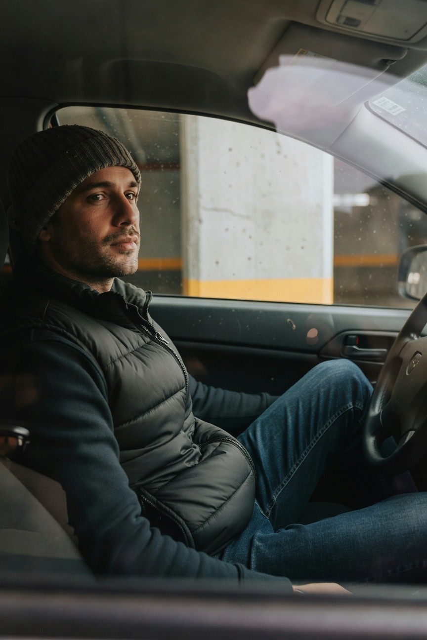 A man sits in the driver's seat of a car in a dimly lit parking garage, wearing a gray beanie, black puffer vest over a dark hoodie, blue jeans, and gazing thoughtfully out the window with one hand on the steering wheel.