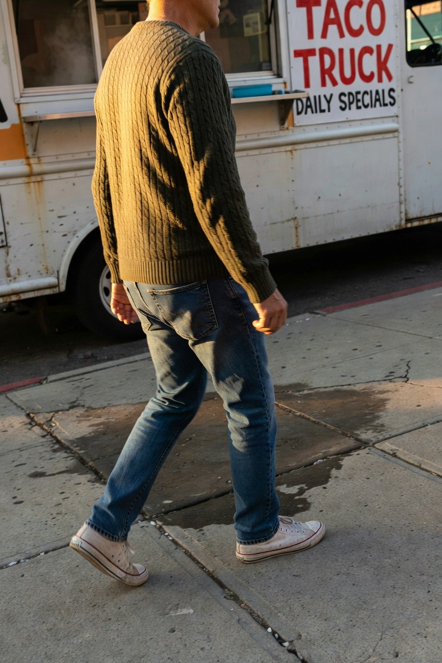 Man in olive green cable knit sweater, faded blue straight-leg jeans, and white low-top Converse sneakers walking past a rusty taco truck on a sunlit urban sidewalk