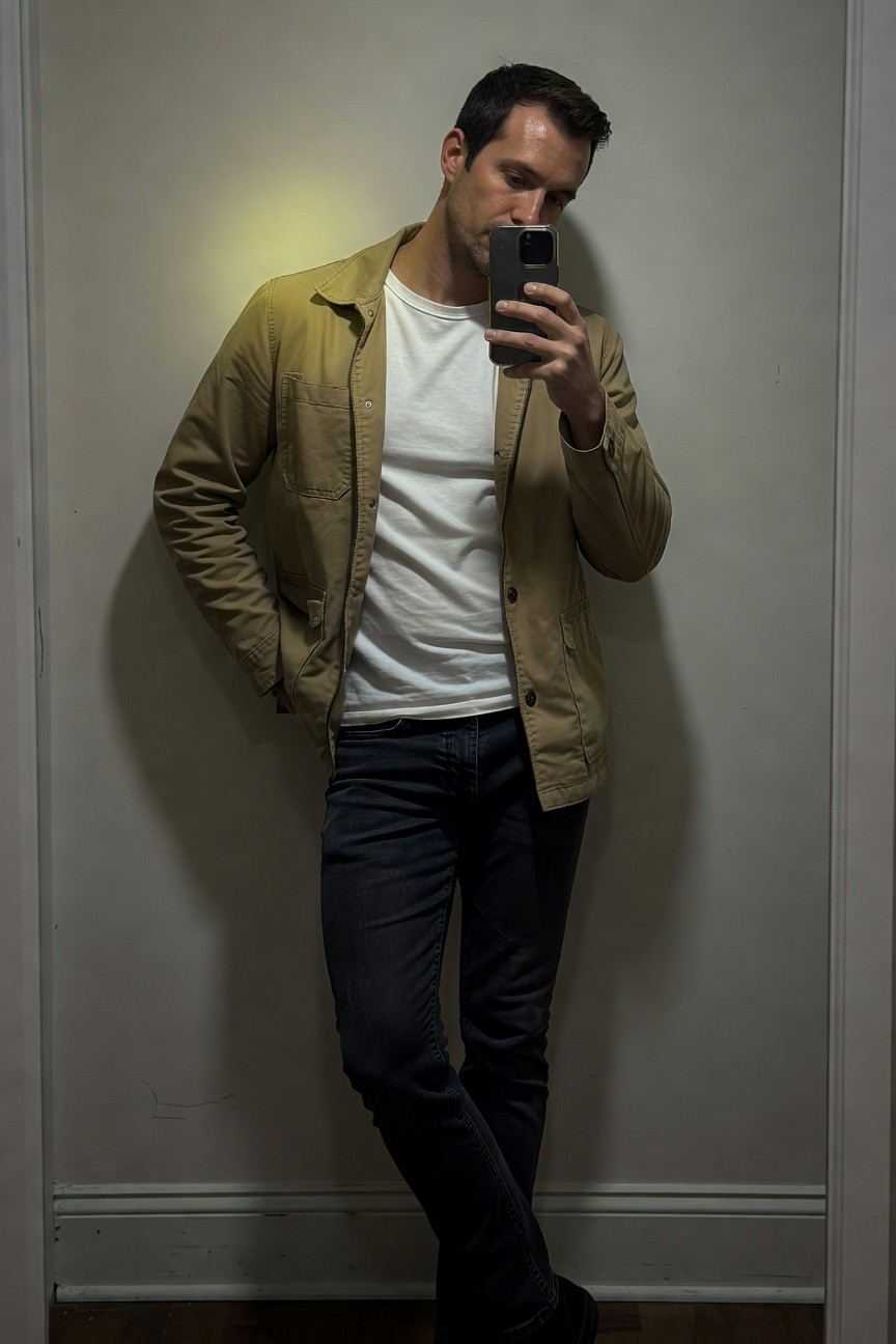 A man stands casually in a beige khaki cotton jacket unbuttoned over a white t-shirt, paired with dark blue straight-leg jeans and black sneakers, taking a mirror selfie in a hallway with one hand in his pocket for a relaxed everyday look