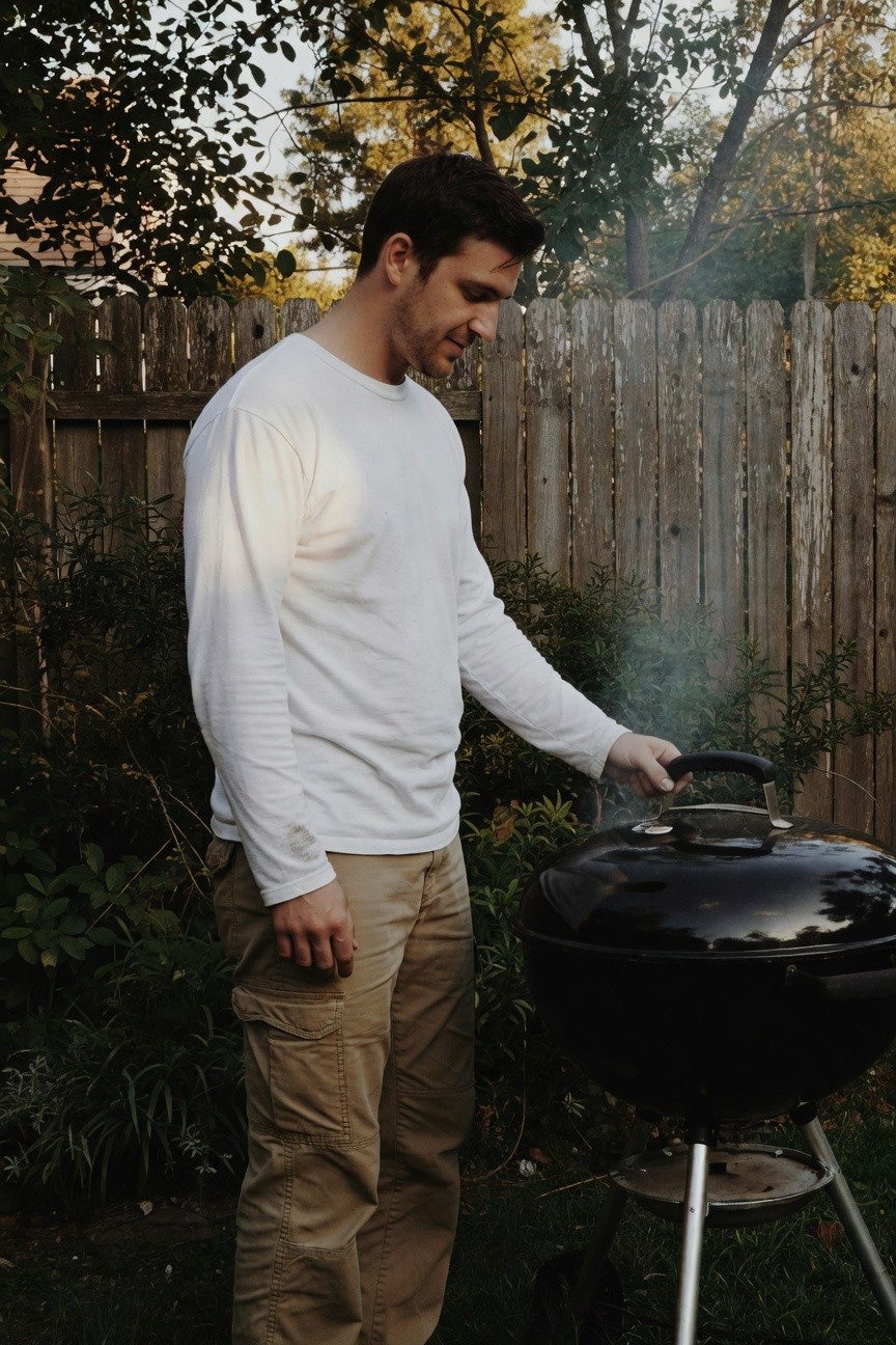 Man in fitted white long-sleeve t-shirt and relaxed khaki cargo pants stands by a smoking black kettle grill in a leafy backyard at dusk, smiling slightly while holding tongs