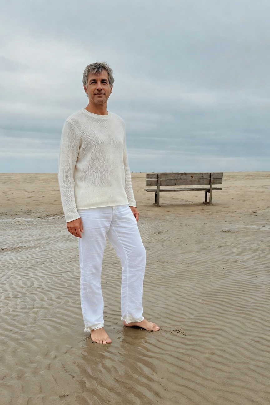 Silver-haired man in cream knit sweater and white wide-leg pants stands barefoot on a misty beach, wooden bench behind him under cloudy skies