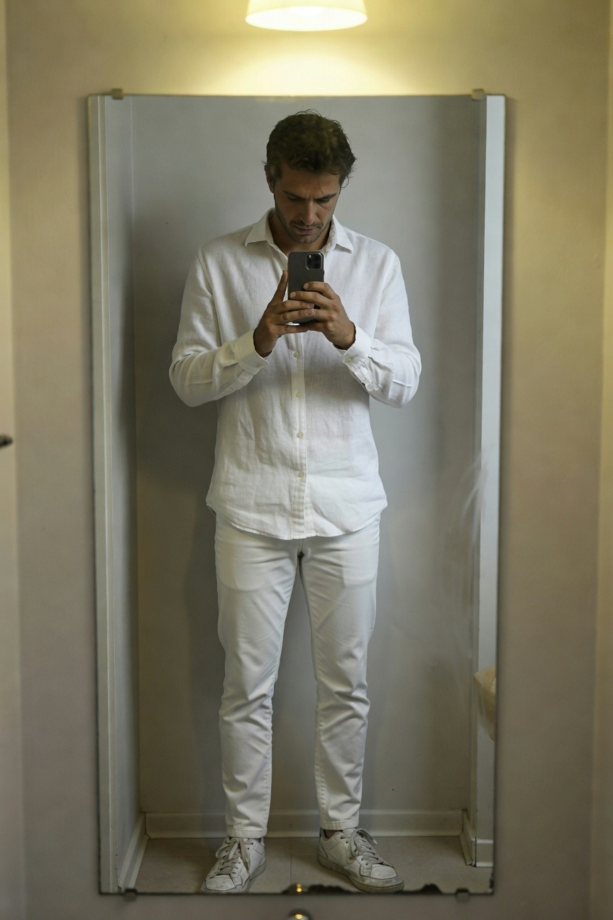 Man in mirror selfie wearing loose-fitting long-sleeve white linen shirt with open collar, slim white trousers, and white sneakers, posed confidently in softly lit room with beige walls
