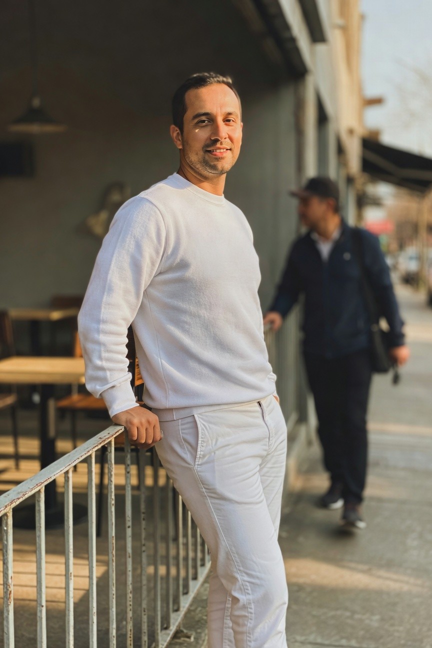 Man in white cable-knit sweater and slim white trousers leaning casually on an outdoor metal railing, smiling confidently in late afternoon urban light with cafe elements blurred behind