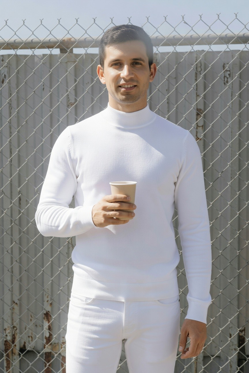 Confident man in fitted white turtleneck sweater and slim white pants stands against rusty chain-link fence holding disposable coffee cup outdoors under hazy sky