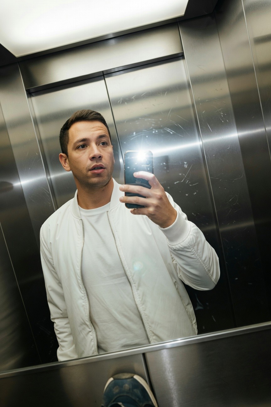 Man in elevator mirror selfie wearing oversized white bomber jacket over white t-shirt, hands in pockets, blue sneakers visible, sleek steel surroundings amplifying the monochrome layers.