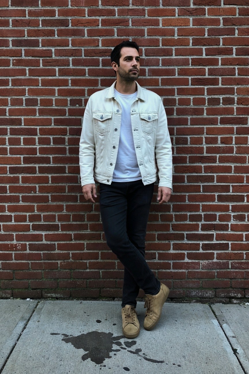 Man leaning casually against red brick wall in light wash white denim jacket over white t-shirt, black slim-fit pants, and beige chunky sneakers, urban street setting with wet pavement