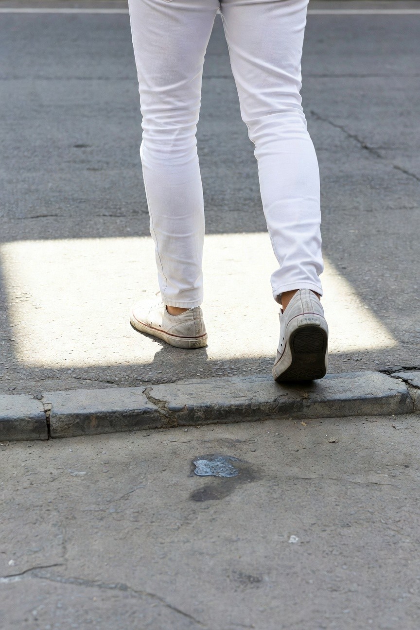 Rear view of a man in slim-fit white jeans and light off-white sneakers walking on a sunlit urban sidewalk with shadows and a small puddle nearby
