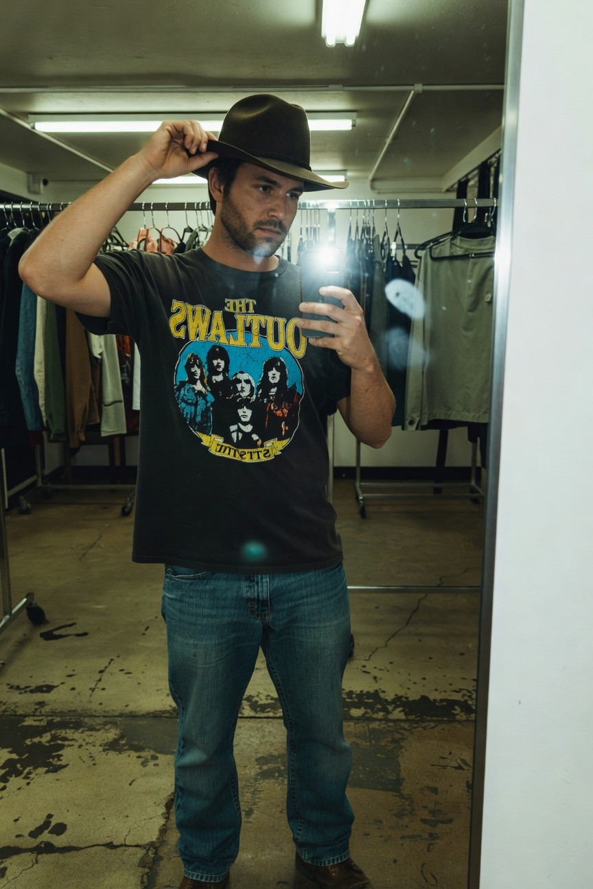 Man in a thrift store mirror selfie wearing a dark gray Outlaws band graphic t-shirt, brown fedora hat, slim blue jeans, and brown boots, holding his phone for the photo