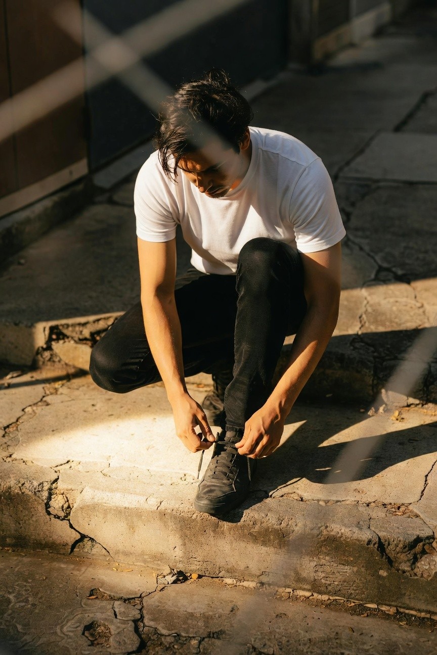 Young man with dark hair in fitted white t-shirt and slim black pants squats on cracked concrete steps in late afternoon sun, tying gray lace-up sneakers, urban alley setting with shadows and light play