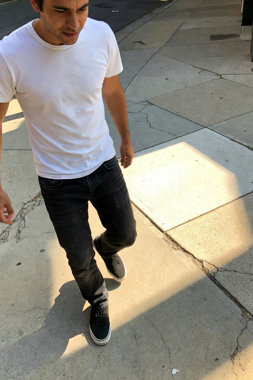 Young man in fitted white cotton t-shirt, slim black jeans, and black low-top sneakers walking on sunny urban sidewalk with concrete and shadows