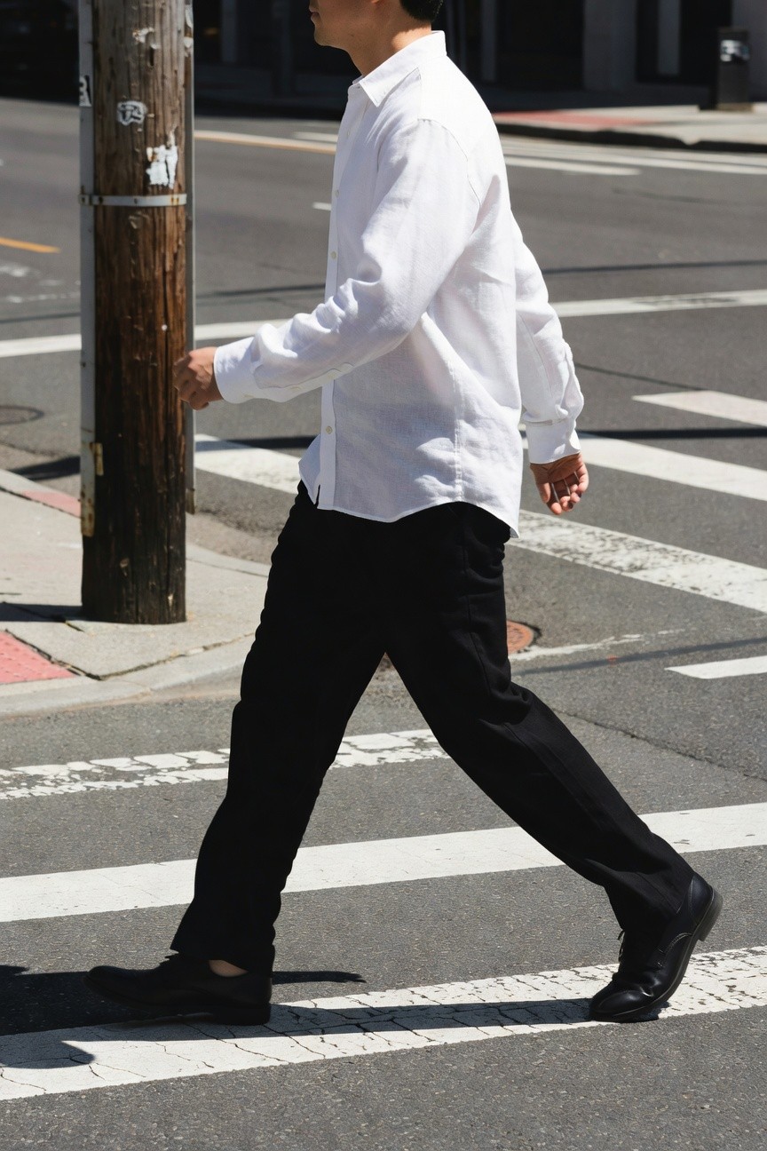 Slim Asian man in untucked white linen button-up shirt and straight-leg black trousers strides across urban crosswalk in black dress shoes, sunlight casting shadows on pavement