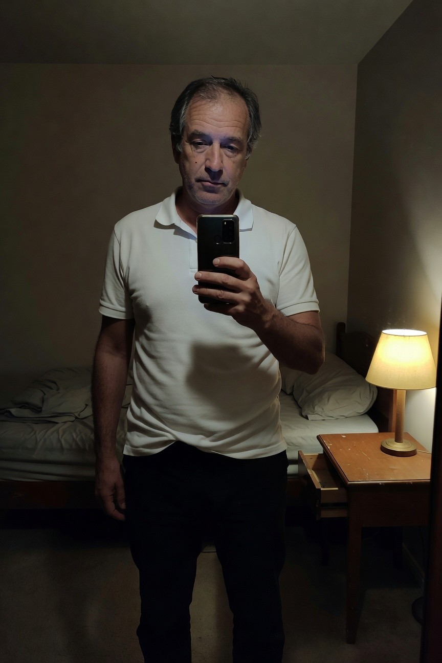 Middle-aged man in a white short-sleeve polo shirt and black pants stands in a dimly lit bedroom taking a selfie, bed and wooden nightstand with lamp in background