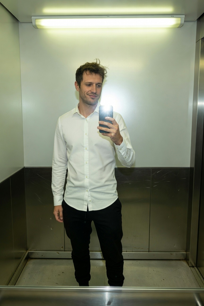 Man in slim white long-sleeve button-up shirt and black slim-fit pants taking a mirror selfie in a modern elevator with fluorescent lighting and steel walls