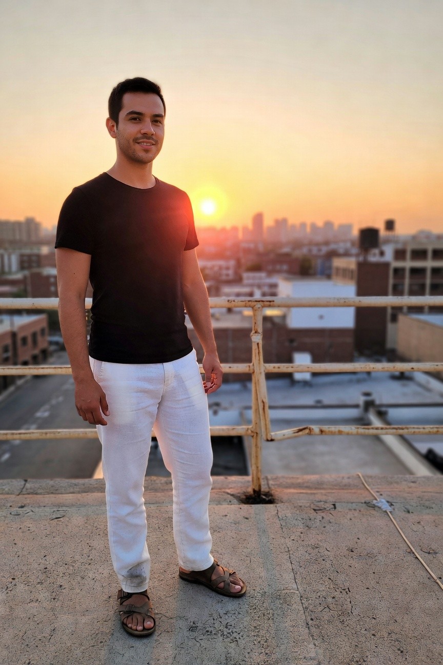 Man in black t-shirt and loose white linen pants stands confidently on urban rooftop at sunset, wearing tan sandals, cityscape and orange sky behind him