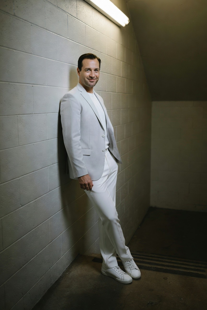Man in light gray blazer, white t-shirt, slim white pants, and white sneakers leaning casually against concrete wall in lit tunnel setting