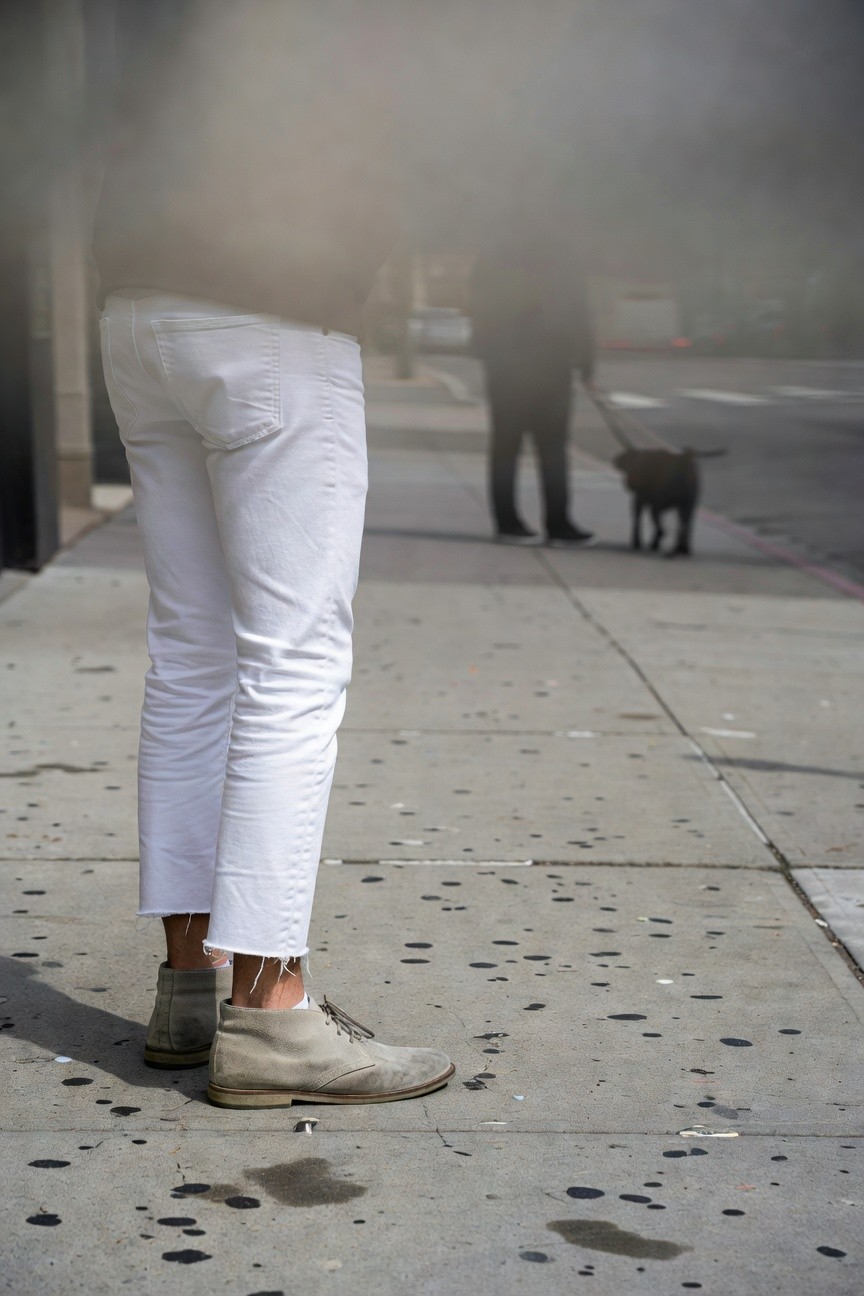 Rear view of a man in slim white frayed-hem jeans and beige suede desert boots, dark jacket, on a foggy urban sidewalk with another figure and small dog nearby